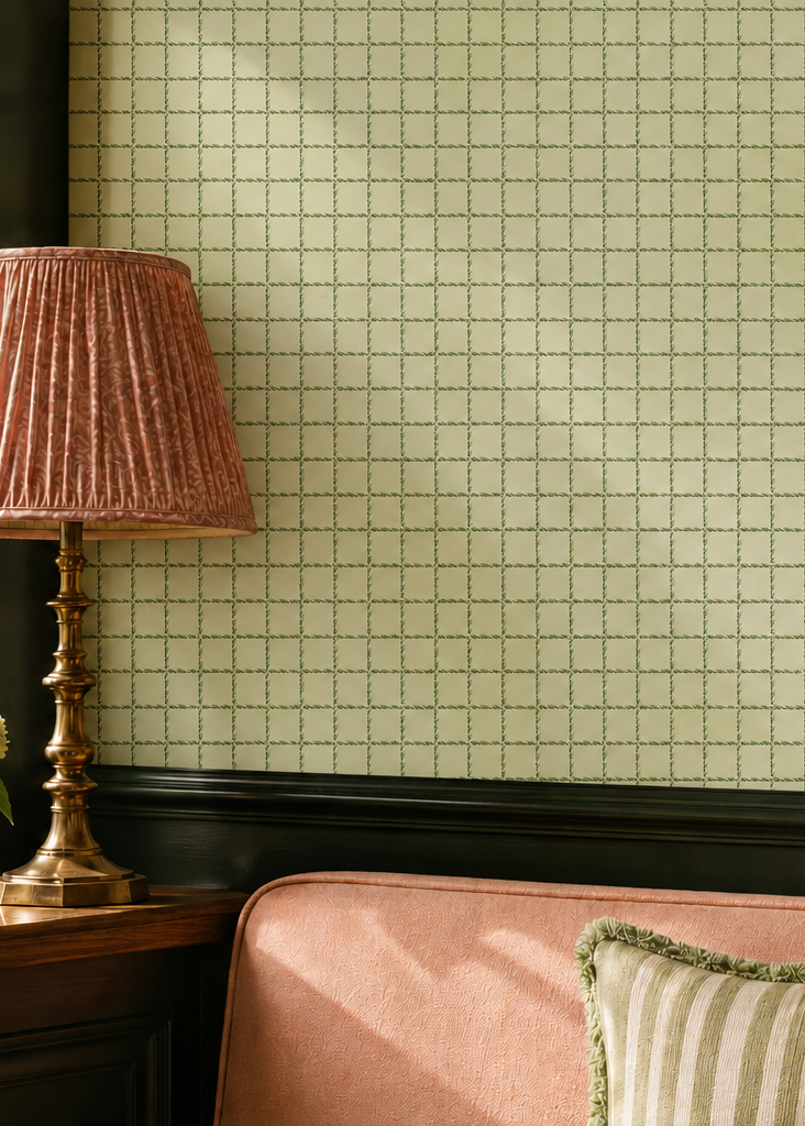 A styled interior scene featuring a pale yellow-green and white grid-patterned wallpaper on a wall above dark wood trim, with a pink armchair, a pleated pink table lamp, a striped pillow, and a vintage tennis racket.