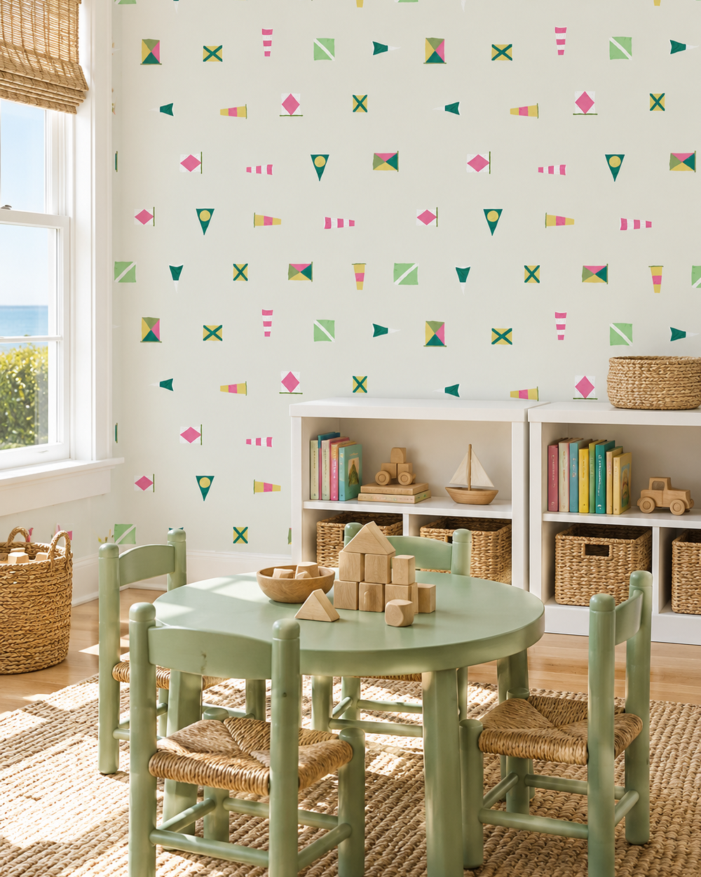 A children's playroom corner featuring a mint green round table and four matching woven-seat chairs, set on a jute rug in front of white shelves with toys. The wall behind is covered in off-white wallpaper decorated with a colorful, repeating pattern of small flags, triangles, and dots. playful and preppy kids nautical flag wallpaper in pink and green for modern lake house nursery in performance vinyl wallpaper
