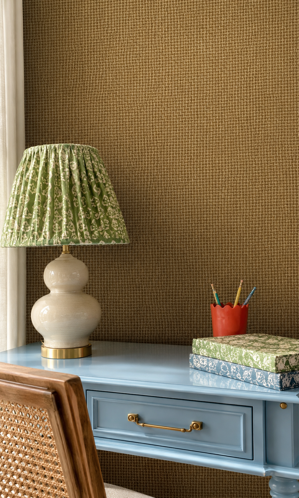A styled interior scene showing a blue desk against a textured brown wall, with a green patterned lampshade, cream gourd table lamp, red pencil cup, patterned books, and part of a woven-back wooden chair visible. Eco-Friendly Tan Basketweave Paperweave Wallpaper – Natural Textural Layers