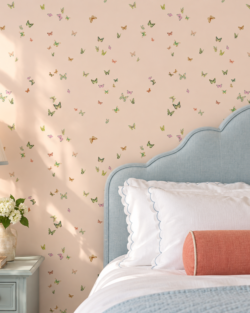 A styled bedroom corner featuring a light blush pink wallpaper with a delicate scattered butterfly pattern, a light blue upholstered headboard, white pillows, and a coral accent pillow. Butterfly Grove wallpaper featuring watercolor-painted butterflies in soft pink, green, and neutral tones inspired by English garden skies perfect for cozy guest bedrooms