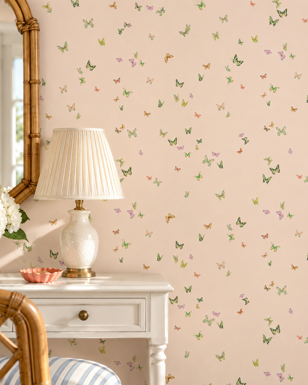 A styled interior corner showing a blush pink wallpaper with a delicate scattered butterfly pattern in green, yellow, and lavender. A white table with a pleated lamp, a bamboo mirror, and a striped cushion are partially visible in the foreground. butterfly grove all-natural and eco-friendly grasscloth and basketweave wallpaper features watercolor-painted butterflies in soft pink, green, and neutral tones inspired by English garden skies perfect for baby girl nurseries 