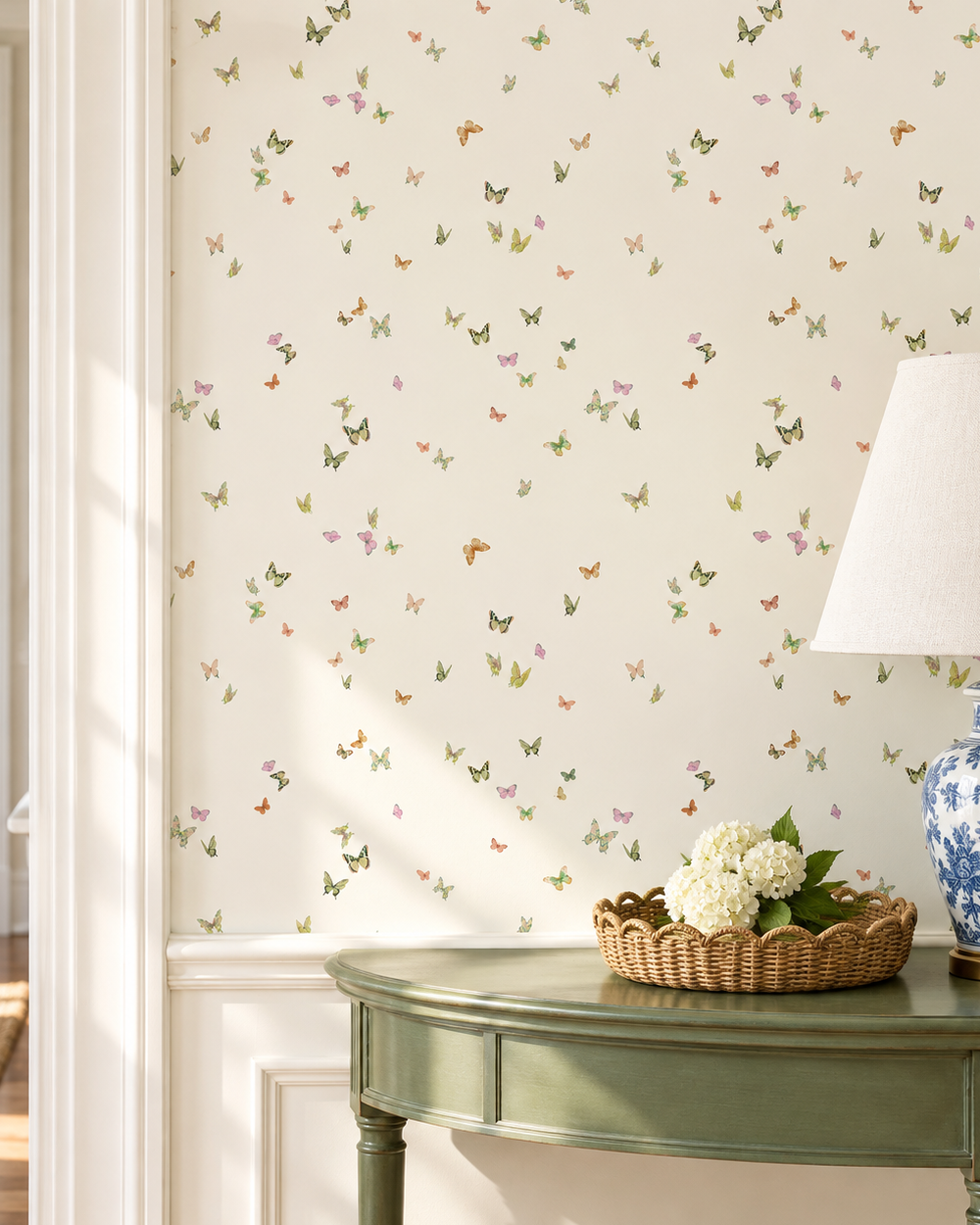 A styled interior scene showing a wall covered in cream wallpaper with a scattered pattern of small multicolored butterflies. A green console table, a blue-and-white ceramic lamp base, and a basket of white flowers are visible in the room. Butterfly Grove wallpaper featuring watercolor-painted butterflies in soft pink, green, and neutral tones inspired by English garden skies printed on a cream colored traditional wall-covering perfect for feminine home offices