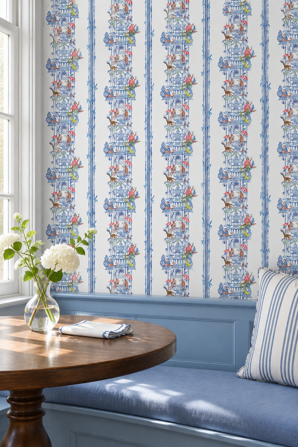 Blue and white patterned wallpaper with vertical stripes, shown in a styled room setting with a blue bench, wooden table, and striped pillow. Hand-painted chinoiserie toile wallpaper on natural grasscloth featuring monkeys, pagodas, bamboo stripes, and whimsical party scenes in multiple colorways.