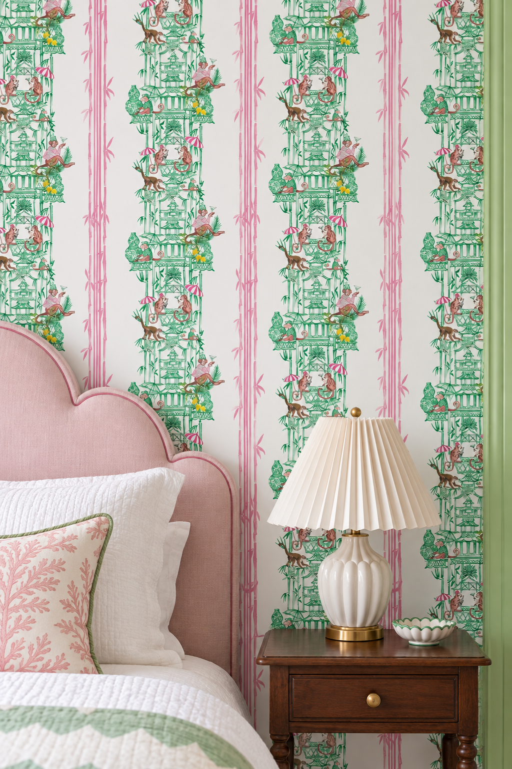 A styled bedroom corner featuring a pink upholstered bed headboard, white bedding, a wooden nightstand with a pleated table lamp, and wallpaper with vertical pink stripes and green toile-style illustrations.