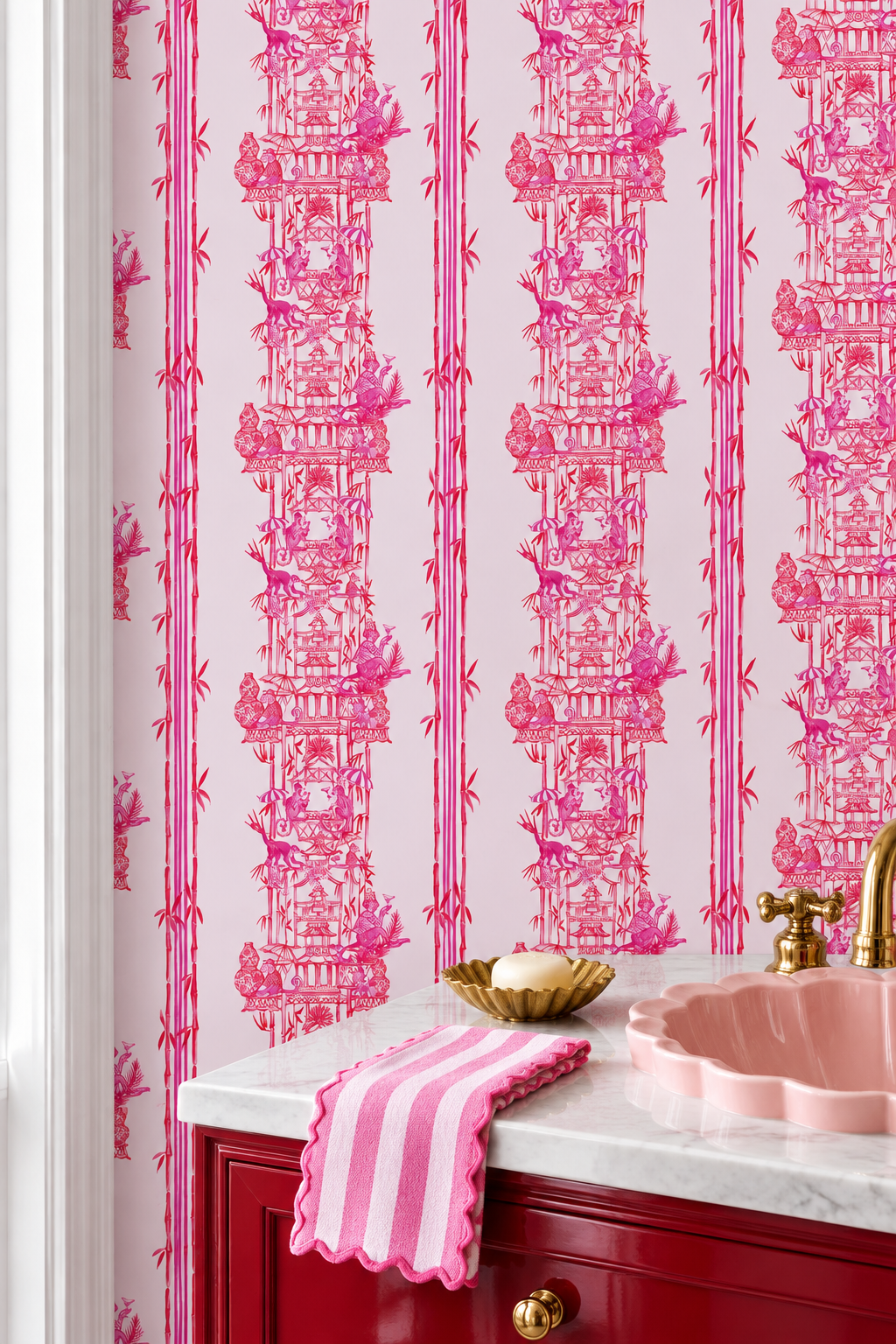Pink and red toile wallpaper with a classic Chinese garden scene pattern, shown behind a red vanity cabinet with a pink sink and a striped pink towel.