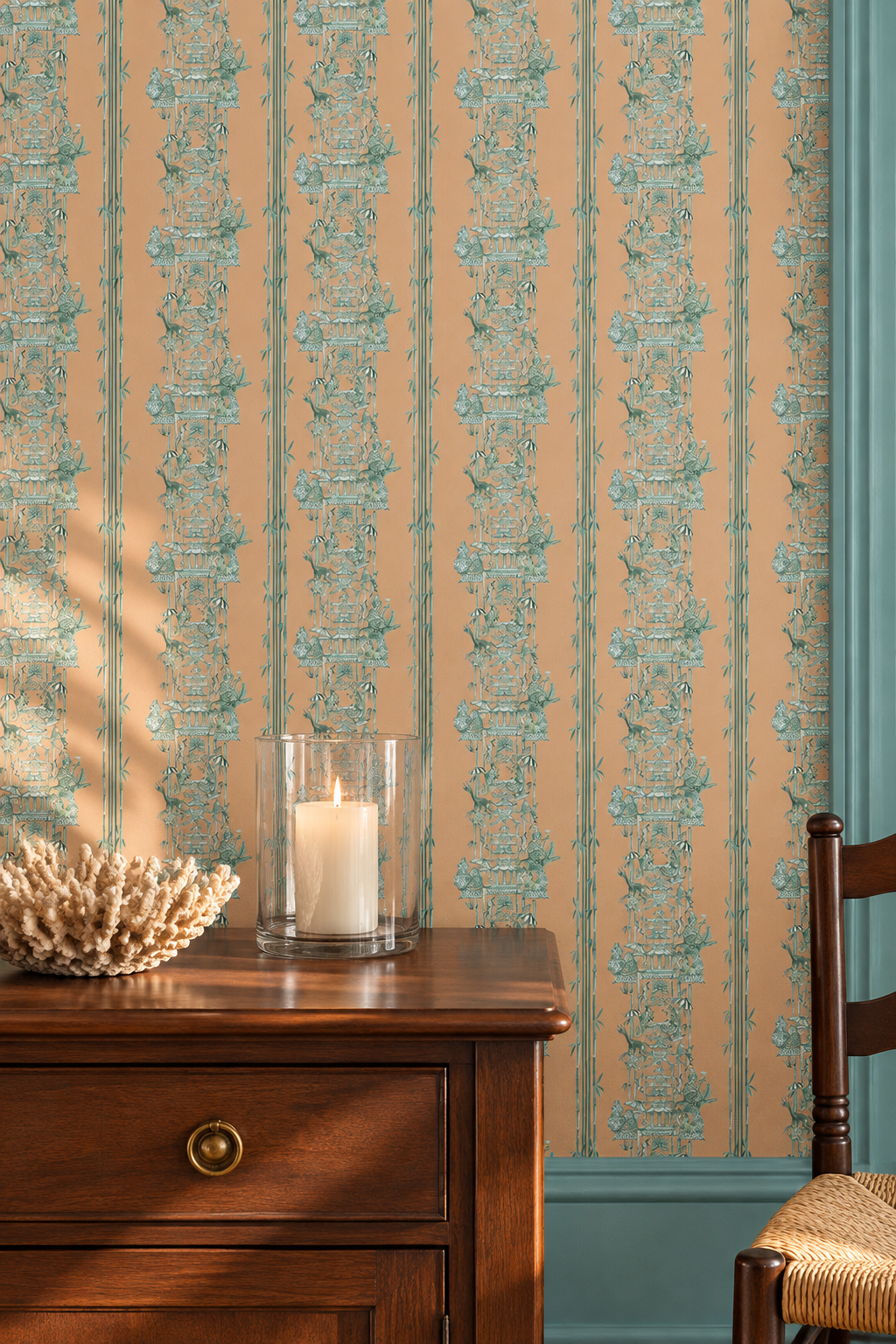 Beige wallpaper with vertical teal-blue ornate stripes, shown in a styled room setting behind a wooden cabinet with a lit candle and a chair partially visible. chinoiserie toile wallpaper, monkey pagoda wallpaper, whimsical chinoiserie wallcovering, bamboo stripe wallpaper, chintz inspired wallpaper, playful toile pattern, hand painted pagoda wallpaper, botanical chinoiserie wallpaper