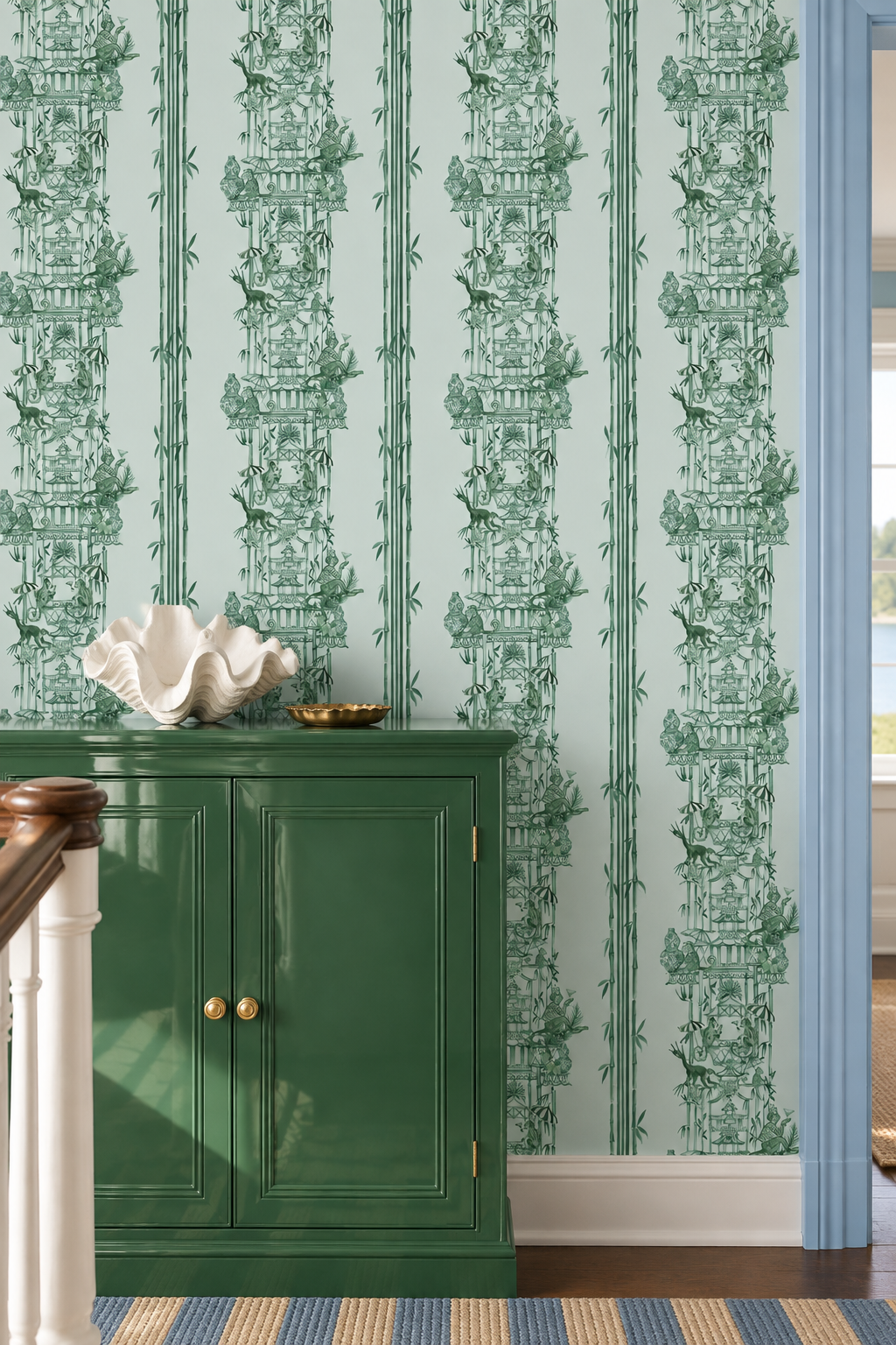 Green toile wallpaper with a vertical pattern of classical figures, birds, and foliage, styled in a room behind a dark green cabinet. Hand-painted chinoiserie toile wallpaper on natural grasscloth featuring monkeys, pagodas, bamboo stripes, and whimsical party scenes in jade green, perfect for luxury foyer