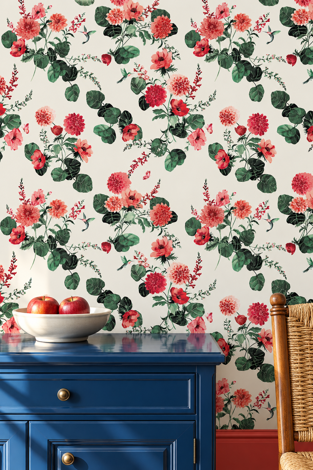 Red and pink floral wallpaper with green leaves on a cream background, shown behind a blue wooden cabinet with a bowl of apples and part of a wicker chair visible. A floral wallpaper pattern featuring large pink and red flowers with green leaves on a white background, shown in a styled interior setting with a wooden table and chairs. chirp society garden wallpaper, hummingbird wallpaper, floral wallpaper, botanical wallpaper, watercolor wallpaper, summer wallpaper, garden wall covering, nature wallpaper, vi