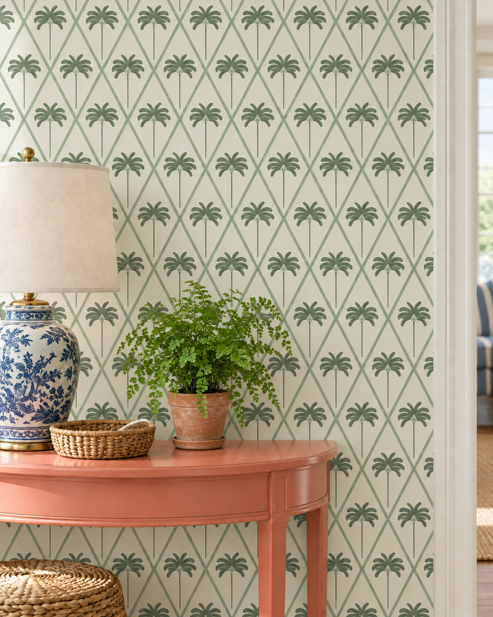 A styled interior scene showing patterned wallpaper with a repeating green palm tree motif on a light background, a coral-pink console table, a blue-and-white ceramic lamp, a potted green plant, and woven baskets. stain and moisture resistant performance vinyl wallpaper, great for wet bars, kitchens and commercial spaces; cream and green botanical ribbon lattice with Palmetto palm trees