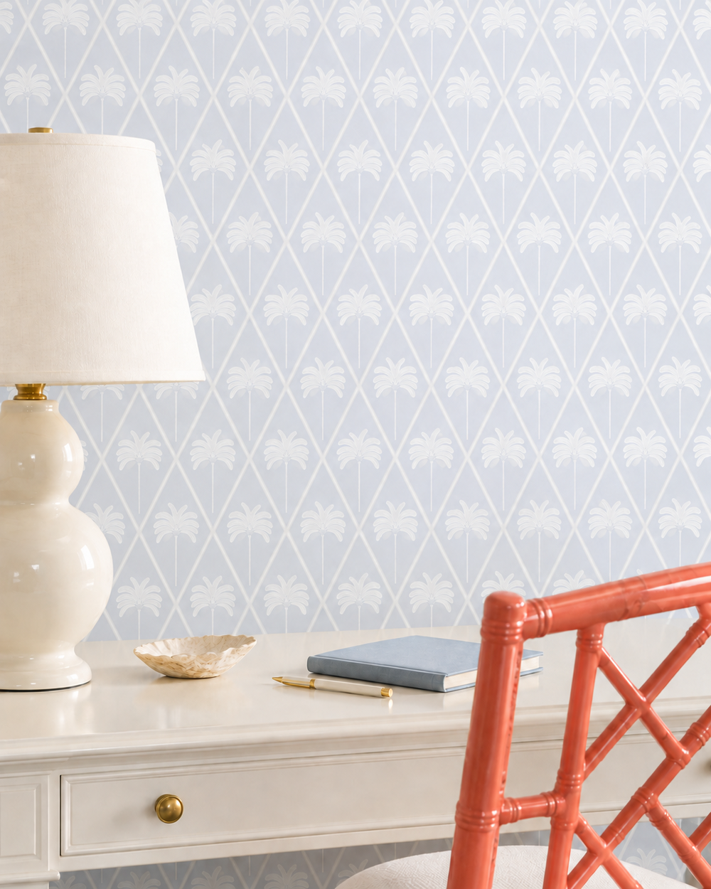 A styled interior scene showing a white desk with a cream table lamp, a small dish, a blue notebook, and a coral-orange chair in front of a pale blue wallpaper patterned with white palm trees and a diamond lattice.