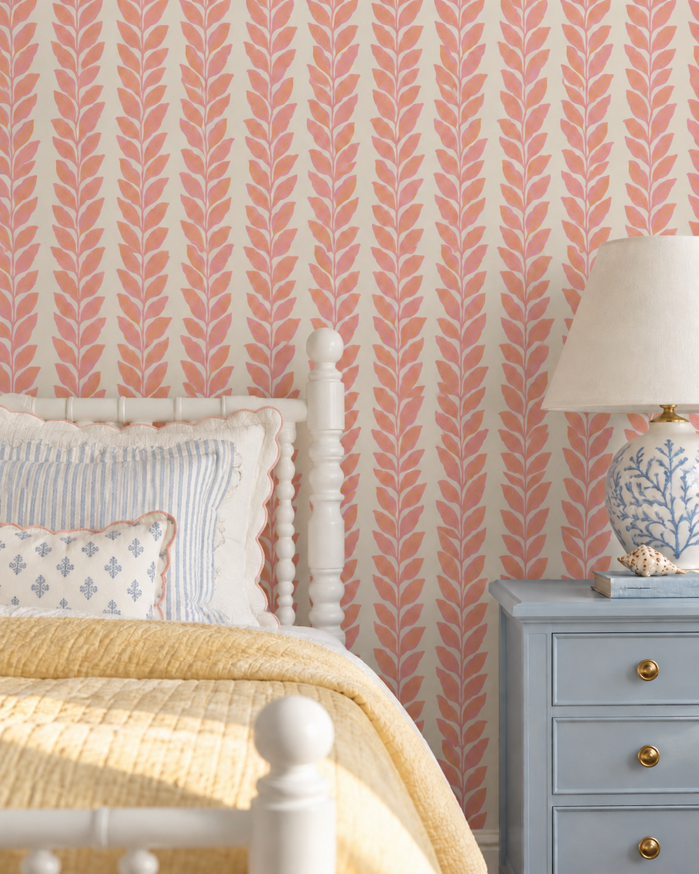 A styled bedroom corner featuring wallpaper with a repeating coral-pink leaf pattern on a warm cream background, a white wooden bed frame, yellow bedding, a light blue nightstand, and a blue-and-white ceramic table lamp.