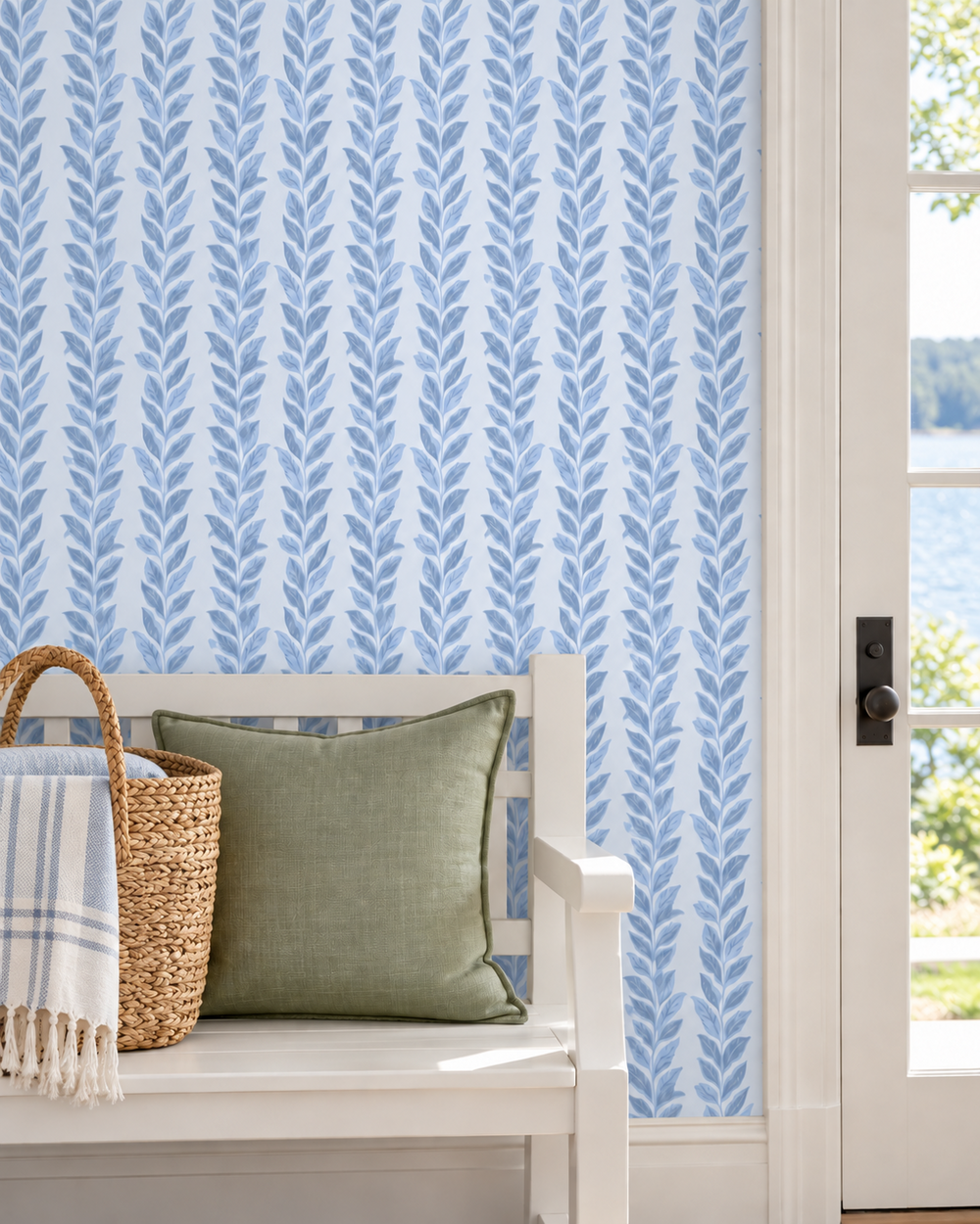 A styled interior corner showing a white bench with a green pillow and woven basket, set against a wall with light blue and white leaf-patterned wallpaper and a white door frame with a view of water outside.