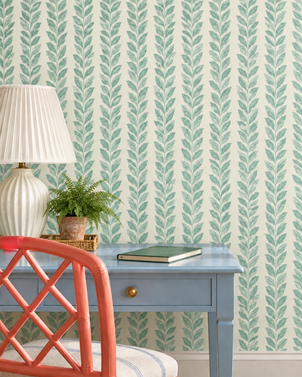 A styled interior scene showing a light blue desk against a wall with repeating vertical leaf-pattern wallpaper in soft green on a warm off-white background. A white pleated lamp, a small potted fern, a closed green book, and part of a coral chair are visible.