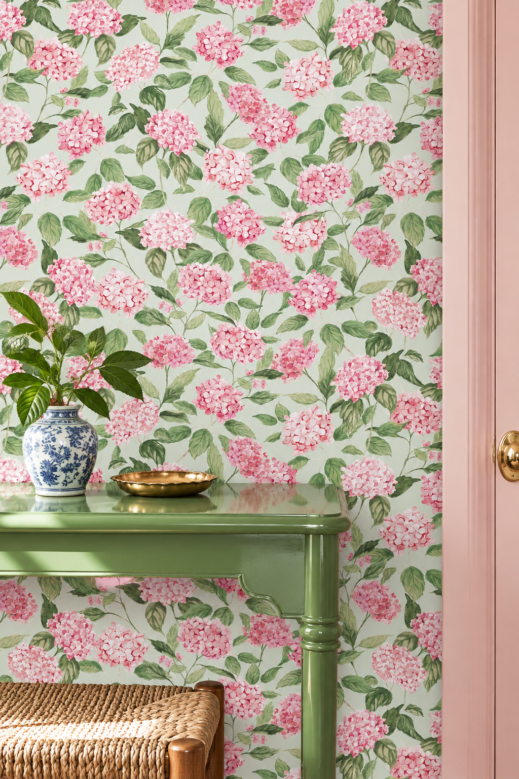 Pink and green floral wallpaper with a repeating hydrangea pattern, shown on a wall behind a green console table with a blue-and-white vase and a gold tray.