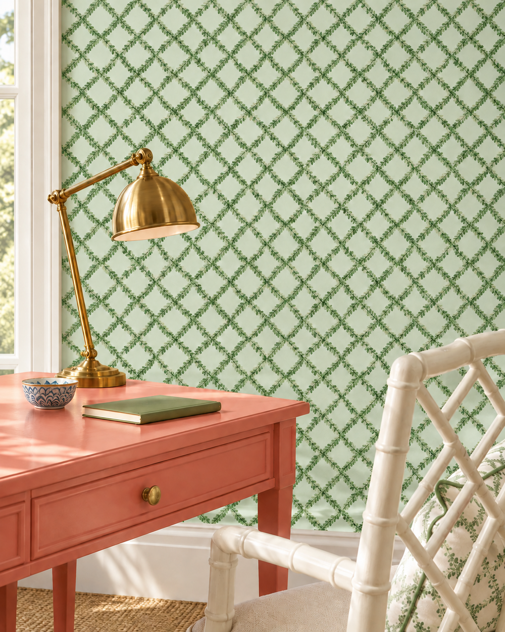 Green and white geometric trellis wallpaper shown in a styled room setting with a coral pink desk, a brass desk lamp, a white bamboo-style chair, and a window.
