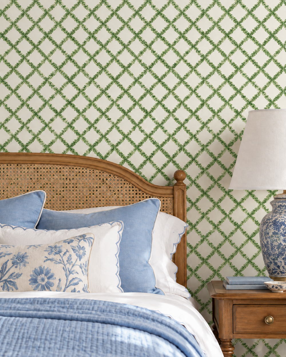 A styled bedroom corner featuring wallpaper with a green trellis pattern on a cream background, a wooden bed with blue and white bedding, and a matching blue-and-white ceramic table lamp on a wooden nightstand.
