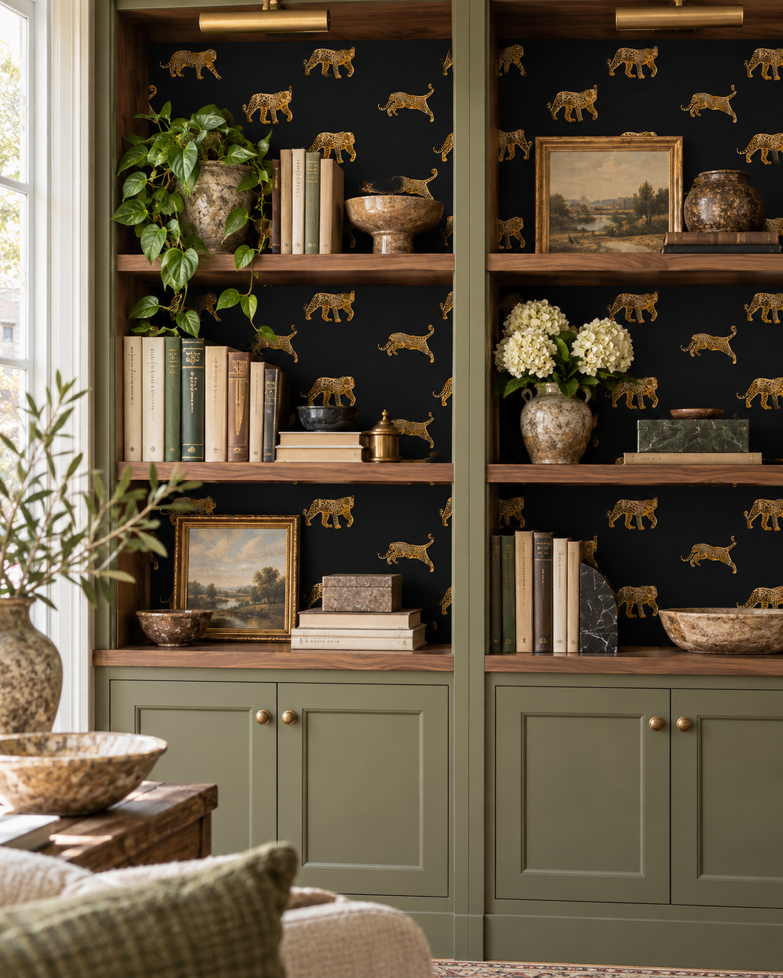 A styled interior scene showing tall built-in shelving with sage green lower cabinets and dark wood shelves. The wall behind is covered in black wallpaper with a repeating gold animal print.