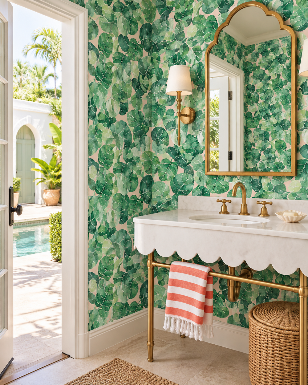 A bathroom interior featuring green tropical leaf pattern wallpaper, a white scalloped vanity with gold hardware, a gold-framed mirror, and an open doorway revealing a poolside outdoor view.
