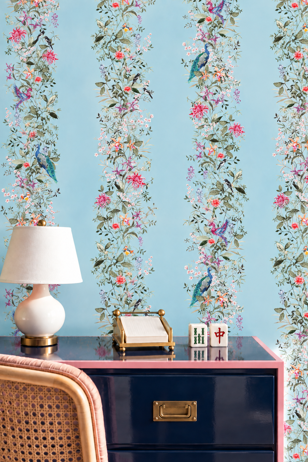 A styled interior scene featuring light blue wallpaper with vertical floral and bird motifs, a white bench with drawers, and two woven rattan chairs on a light rug. Natural grasscloth wallpaper featuring hand-painted Mahjong Garden Stripe with chinoiserie florals, peacocks, birds, bamboo, and botanical watercolor artwork.