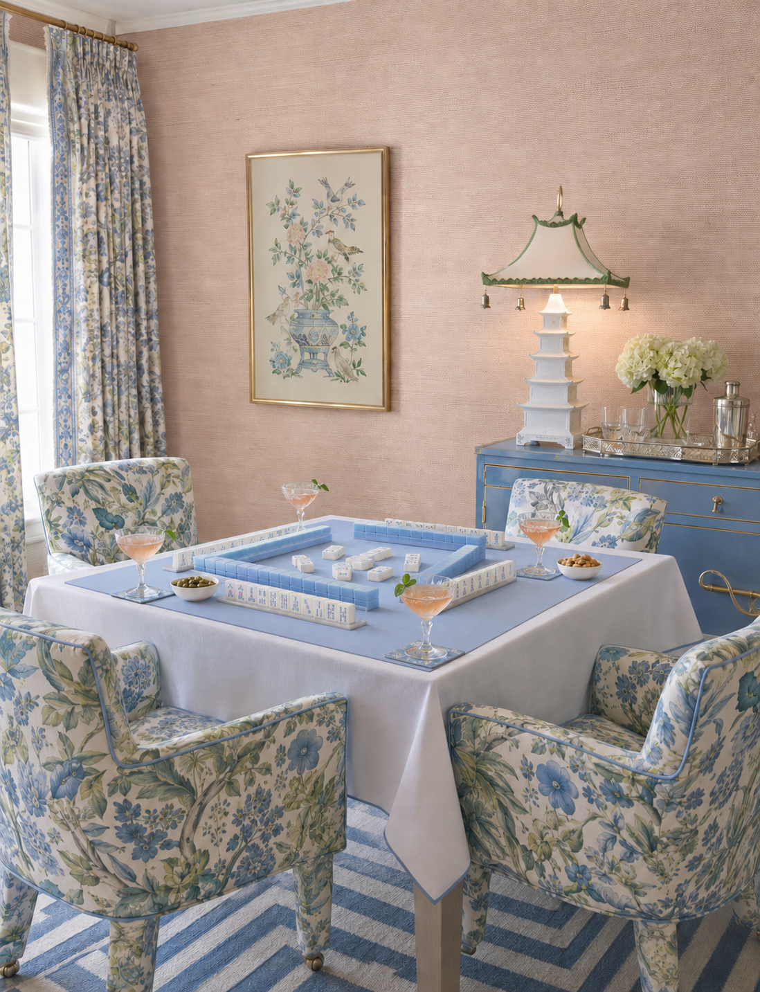 A styled dining room scene with a blue-and-white floral upholstered dining table set with four matching armchairs, a white tablecloth, cocktail glasses, a blue sideboard, and a pink wall with floral artwork and light pink blush solid dyed sisal wallpaper