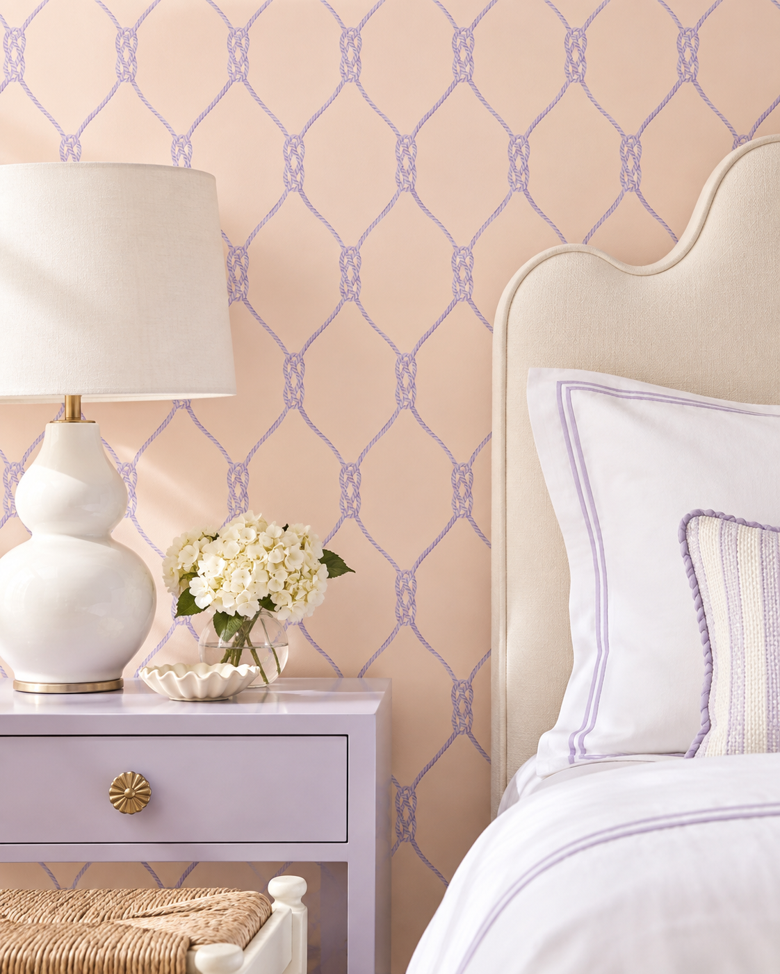 A styled bedroom corner featuring lavender and cream bedding, a matching lavender nightstand, a white ceramic table lamp, and a decorative vase of white flowers against a peach wallpaper with a light purple trellis pattern. Matte wallpaper featuring a repeating twisted rope-knot geometric pattern on a smooth traditional paper surface on neutral pink 