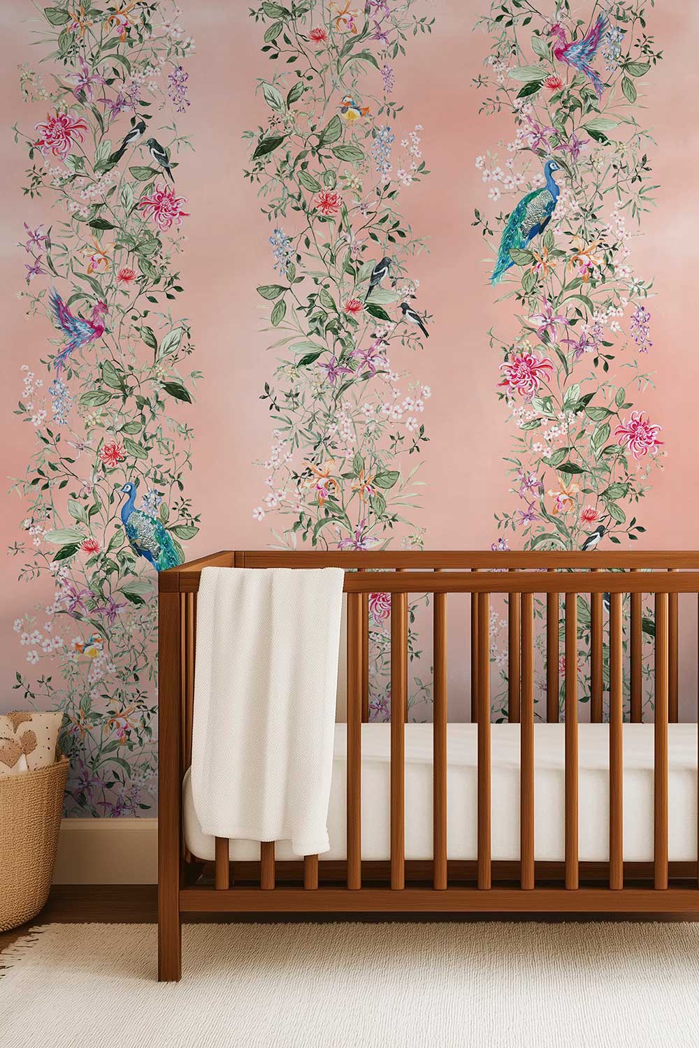 A wooden baby crib with a white blanket in a nursery setting, set against a pink wallpaper decorated with vertical floral vines and blue birds. Hand-painted Mahjong Garden Stripe wallpaper featuring watercolor chinoiserie florals, peacocks, birds, bamboo, and botanical stripes on an blush pink coral background