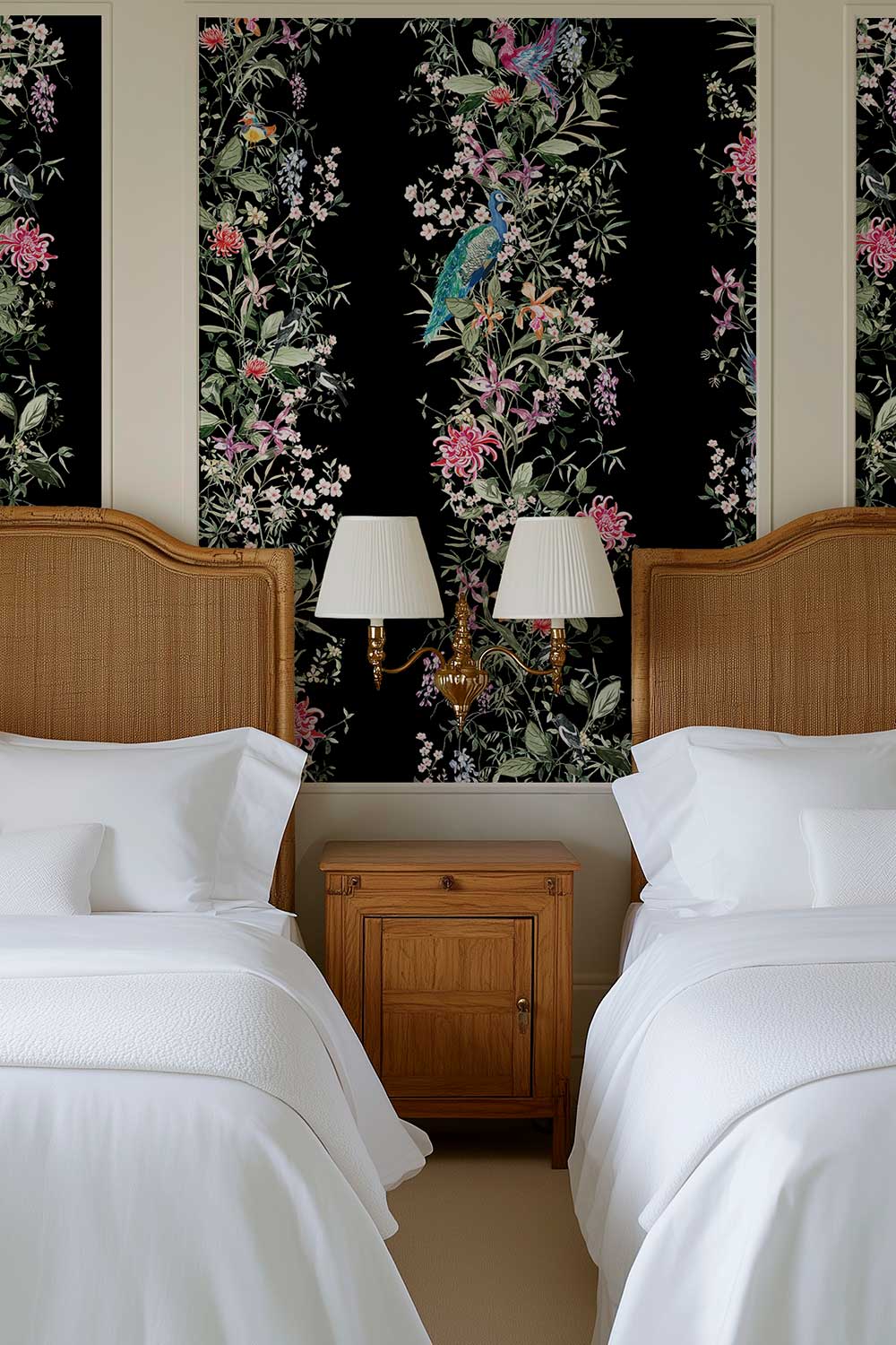 A styled bedroom interior with two twin beds dressed in white bedding, wooden headboards, a small wooden nightstand, and two wall-mounted lamps against a black floral wallpaper featuring a peacock and botanical pattern. Natural grasscloth wallpaper featuring hand-painted Mahjong Garden Stripe with chinoiserie florals, peacocks, birds, bamboo, and botanical watercolor artwork.