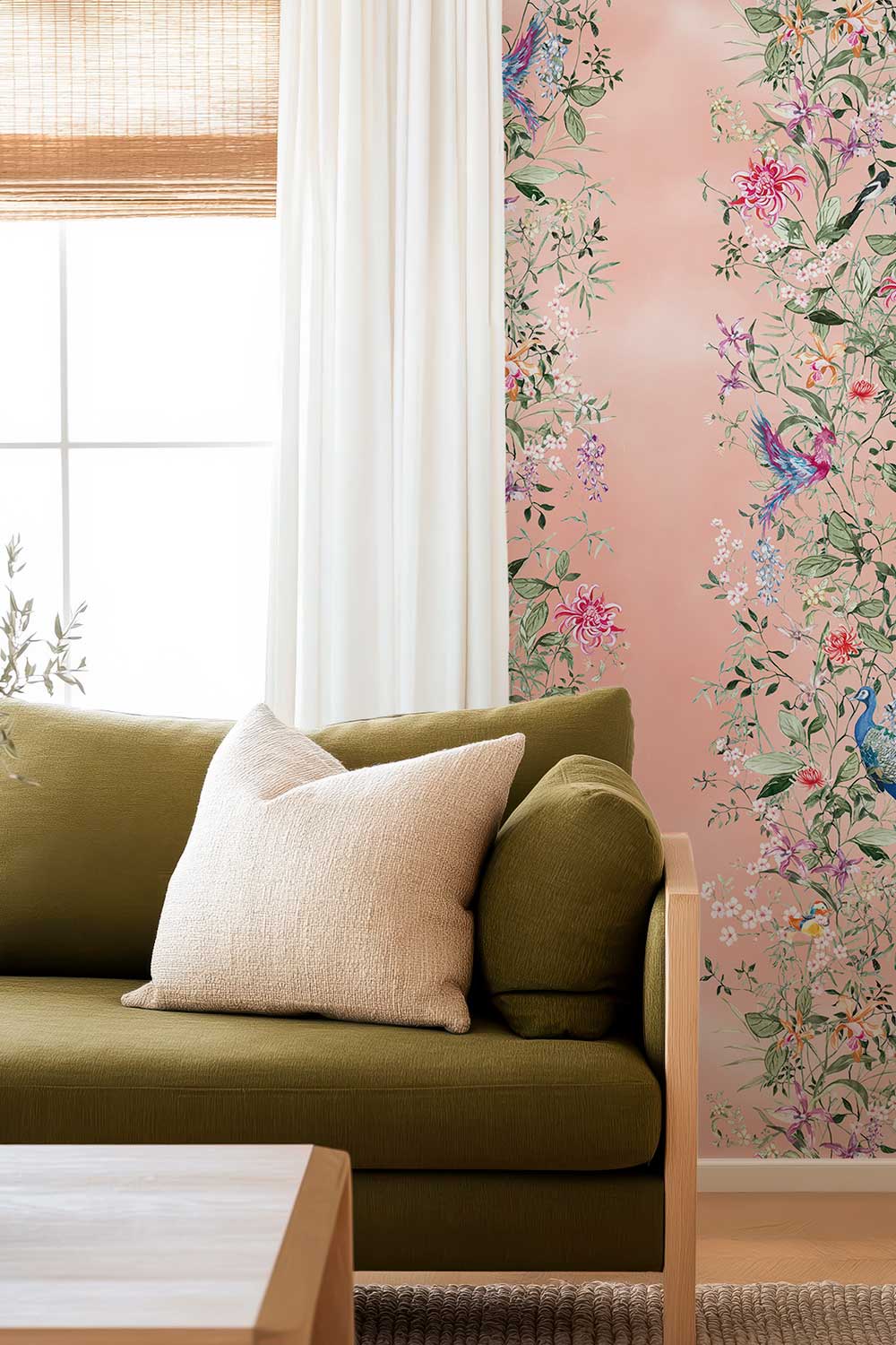 An olive green sofa with a cream throw pillow in a bright living room, set against a wall featuring pink floral and bird wallpaper. Natural grasscloth wallpaper featuring hand-painted Mahjong Garden Stripe with chinoiserie florals, peacocks, birds, bamboo, and botanical watercolor artwork.