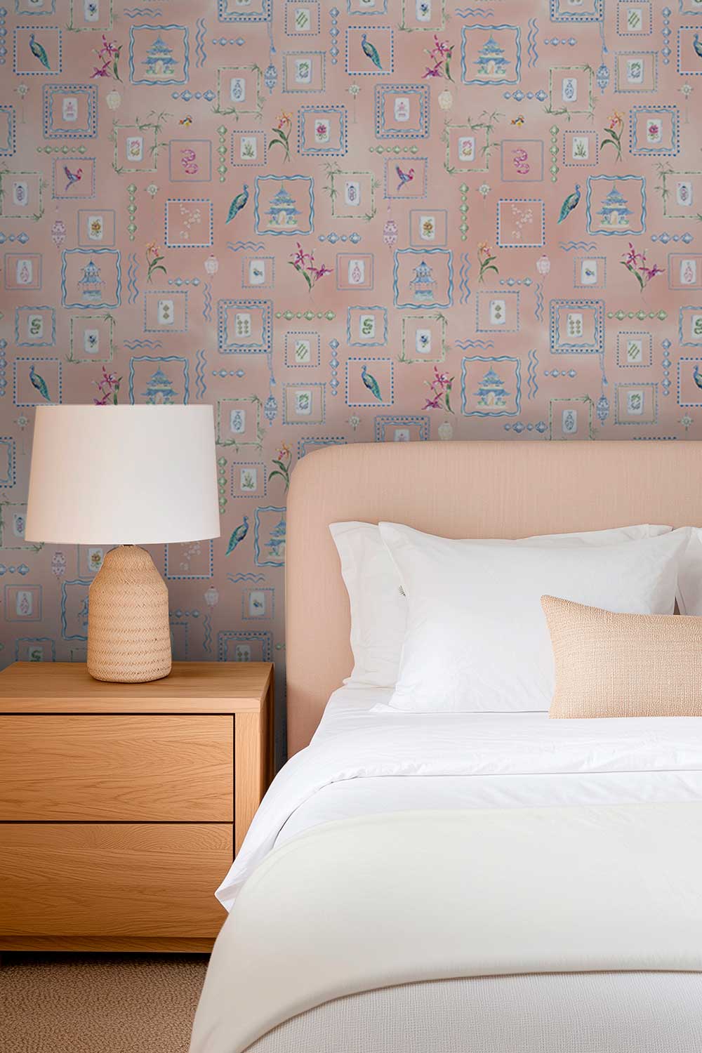 A styled bedroom corner featuring a bed with white linens and a beige pillow, a wooden nightstand with a textured white lamp, and a pink patterned wallpaper background. Hand-painted watercolor Mahjong Tiles wallpaper featuring mahjong tiles, pagodas, bamboo, birds, ginger jars, lanterns, and chinoiserie artwork on a soft pink coral background printed on an all-natural and eco-friendly grasscloth wall-covering.