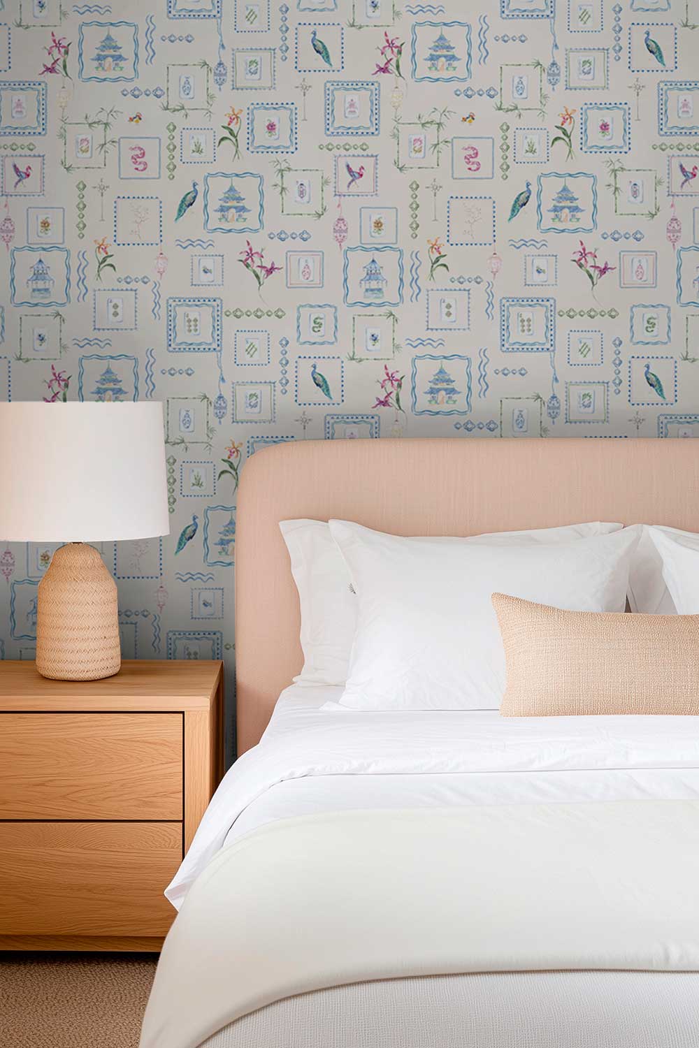 A styled bedroom scene showing a neatly made bed with white bedding and a light peach upholstered headboard, set against a patterned wallpaper with blue, pink, and green motifs. A wooden nightstand with a white lamp is visible on the left. Hand-painted watercolor Mahjong Tiles wallpaper featuring mahjong tiles, pagodas, bamboo, birds, ginger jars, lanterns, and chinoiserie artwork on an off-white background printed on an all-natural and eco-friendly grasscloth wall-covering.