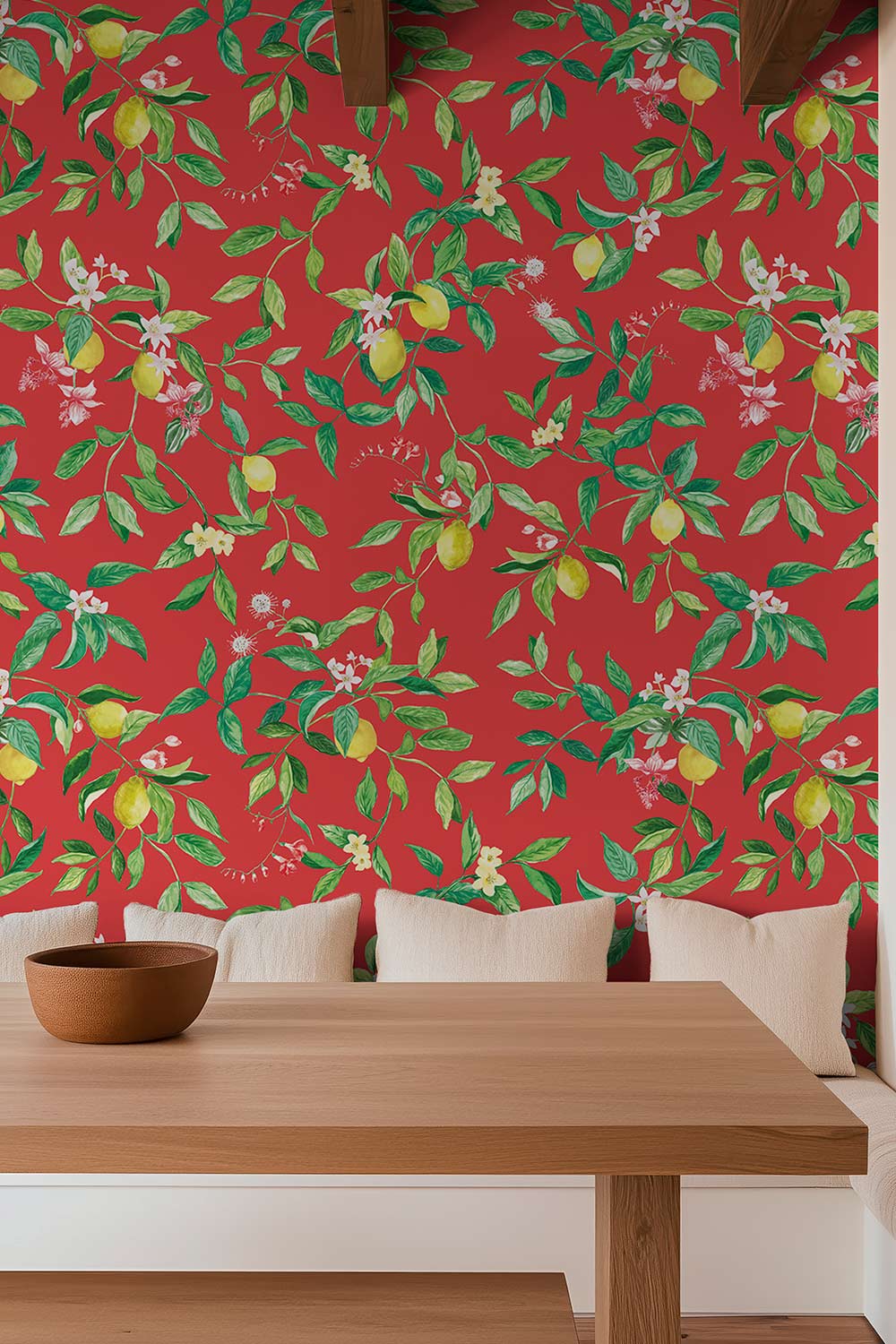 A red wallpaper with a green botanical pattern of lemons, branches, and small white flowers, shown behind a light wood dining table and white pillows. lemon wallpaper, citrus wallpaper, limoncello wallpaper, white and pastel wallpaper, fruit wallpaper, italian inspired wallpaper, kitchen wallpaper, lemon floral print on all-natural and eco-friendly wall-covering grasscloth and paperweave