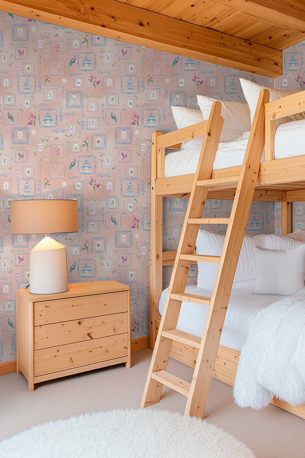 A cozy children's bedroom featuring a light natural wood bunk bed with white bedding, a matching wooden three-drawer nightstand with a table lamp, and pink patterned wallpaper. Hand-painted watercolor Mahjong Tiles wallpaper featuring mahjong tiles, pagodas, bamboo, birds, ginger jars, lanterns, and chinoiserie artwork on a soft pink coral background printed on an all-natural and eco-friendly grasscloth wall-covering.