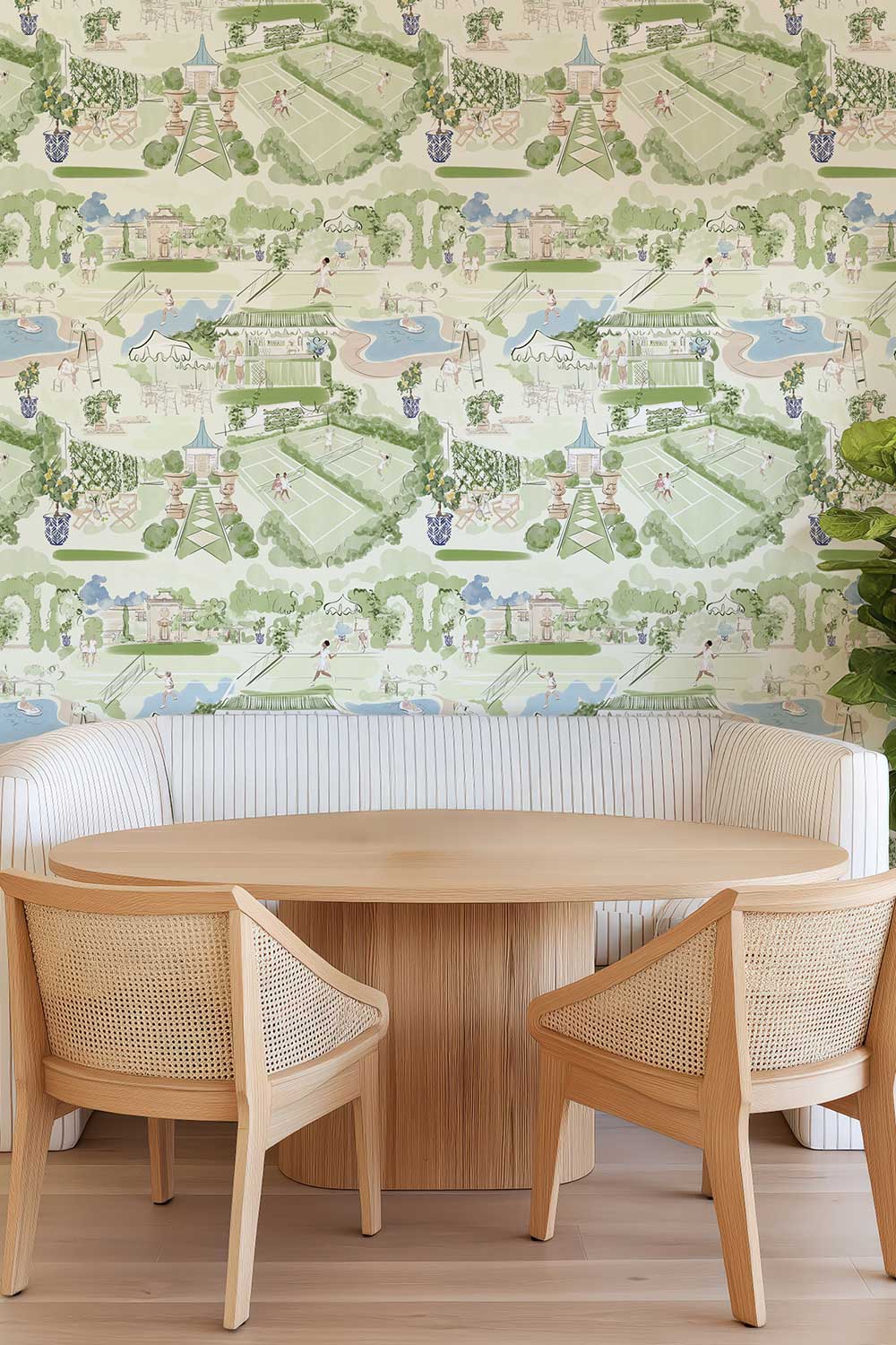 A dining area featuring a round light wood table and two matching wooden chairs with woven cane seats, set in front of a white upholstered banquette. The wall is covered in a detailed pastel landscape wallpaper depicting a luxury tennis club. Tennis Club Toile removable peel and stick wallpaper featuring watercolor tennis players, green courts, racquets, a swimming pool and elegant clubhouse gardens.