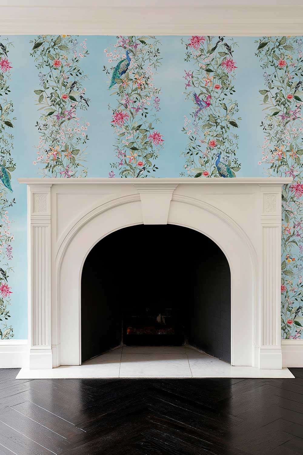 A white arched fireplace mantel set against a light blue wallpaper with vertical floral and bird motifs, above a dark herringbone wood floor. Hand-painted Mahjong Garden Stripe wallpaper featuring watercolor chinoiserie florals, peacocks, birds, bamboo, and botanical stripes on a soft blue background