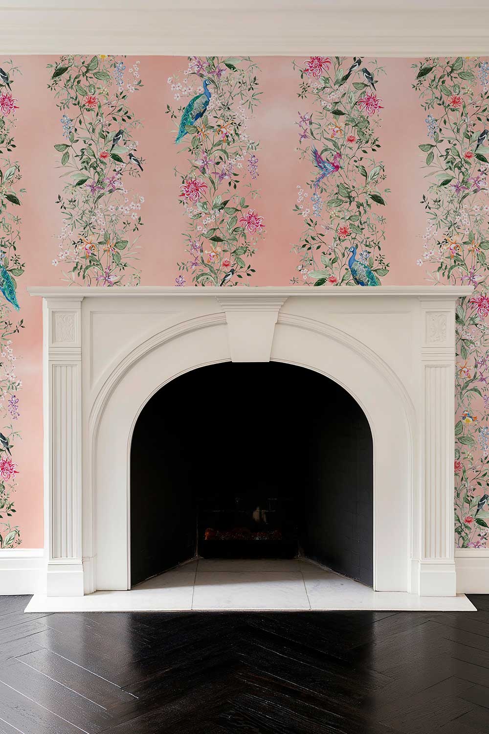 A white arched fireplace mantel set against pink floral wallpaper with peacock motifs, above a dark herringbone wood floor. Hand-painted Mahjong Garden Stripe wallpaper featuring watercolor chinoiserie florals, peacocks, birds, bamboo, and botanical stripes on an blush pink coral background