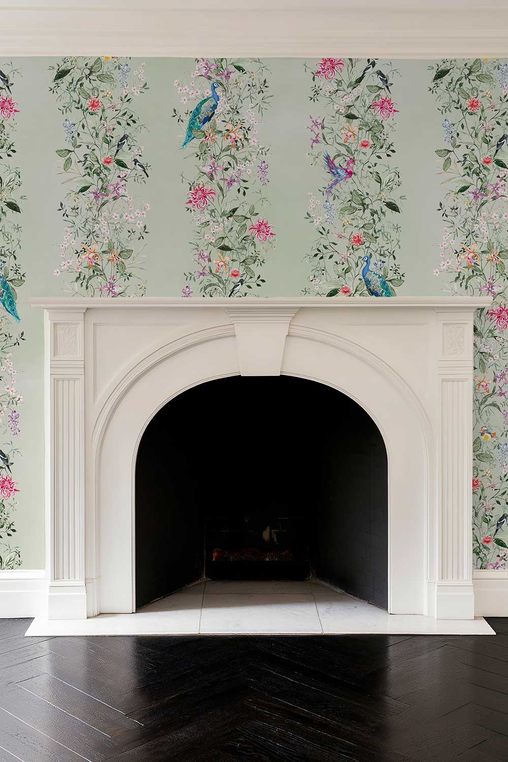 A white arched fireplace mantel set against wallpaper with vertical floral and bird motifs on a pale green background, above a dark wood floor. Natural grasscloth wallpaper featuring hand-painted Mahjong Garden Stripe with chinoiserie florals, peacocks, birds, bamboo, and botanical watercolor artwork.
