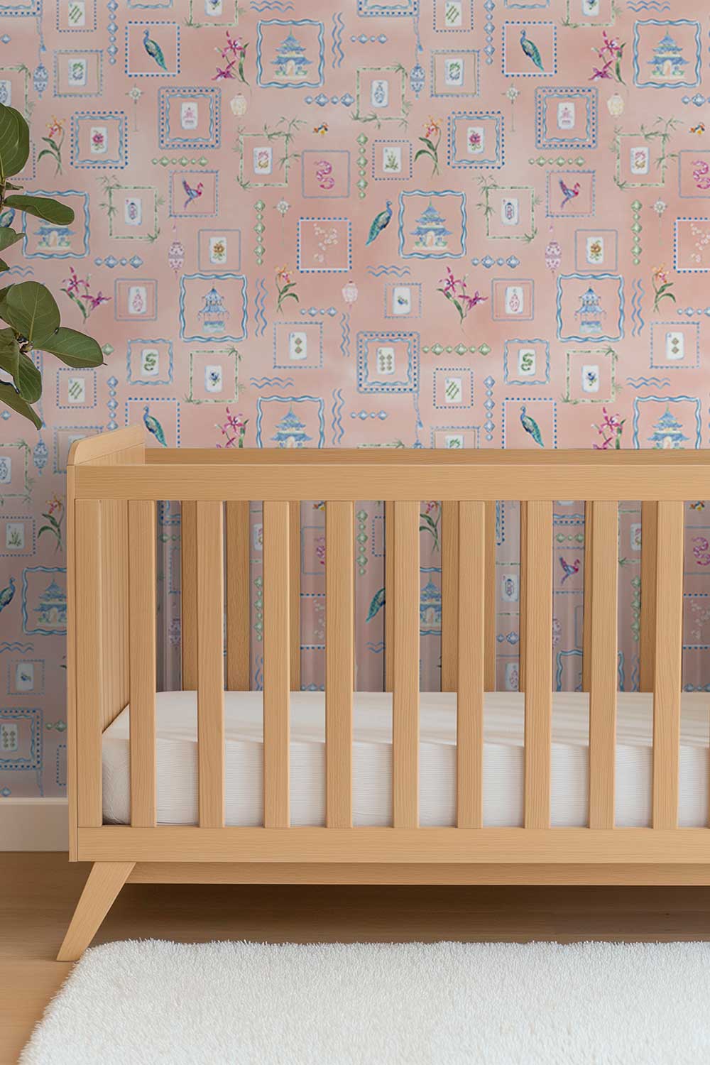 A light natural wood crib with vertical slats and angled legs, shown in a nursery setting against a pink patterned wallpaper background with a white rug in the foreground. Washable performance vinyl Mahjong Tiles wallpaper with hand-painted chinoiserie details, perfect for powder rooms, bathrooms, game rooms, and clubs.