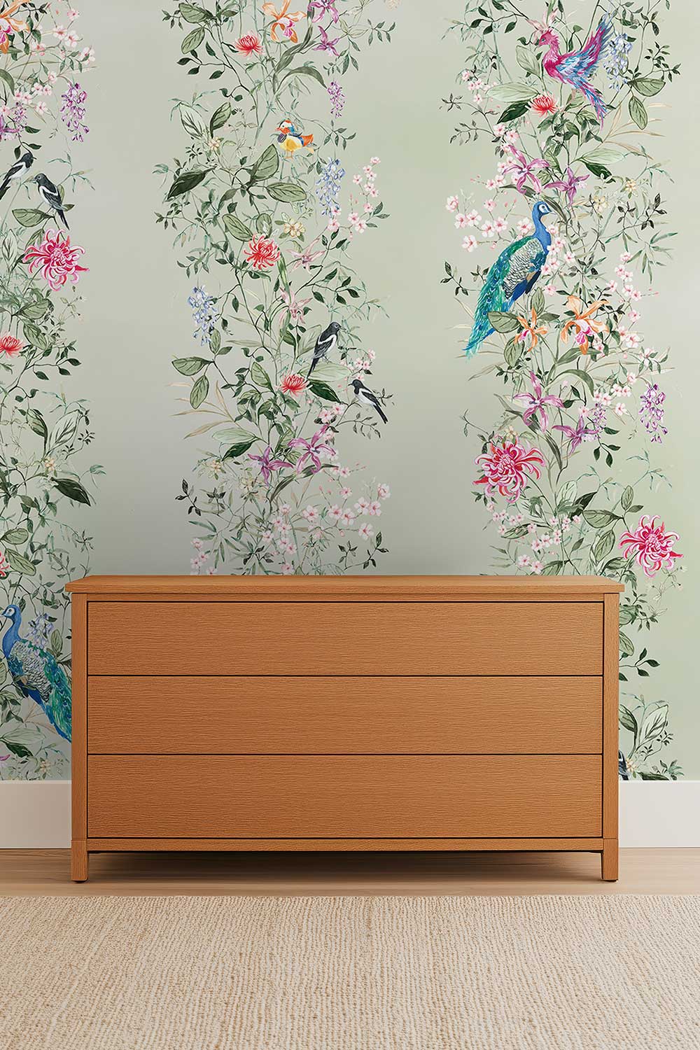A light wood three-drawer dresser placed against a pale green wall with a colorful floral and bird wallpaper pattern. Hand-painted Mahjong Garden Stripe wallpaper featuring watercolor chinoiserie florals, peacocks, birds, bamboo, and botanical stripes on a soft green background