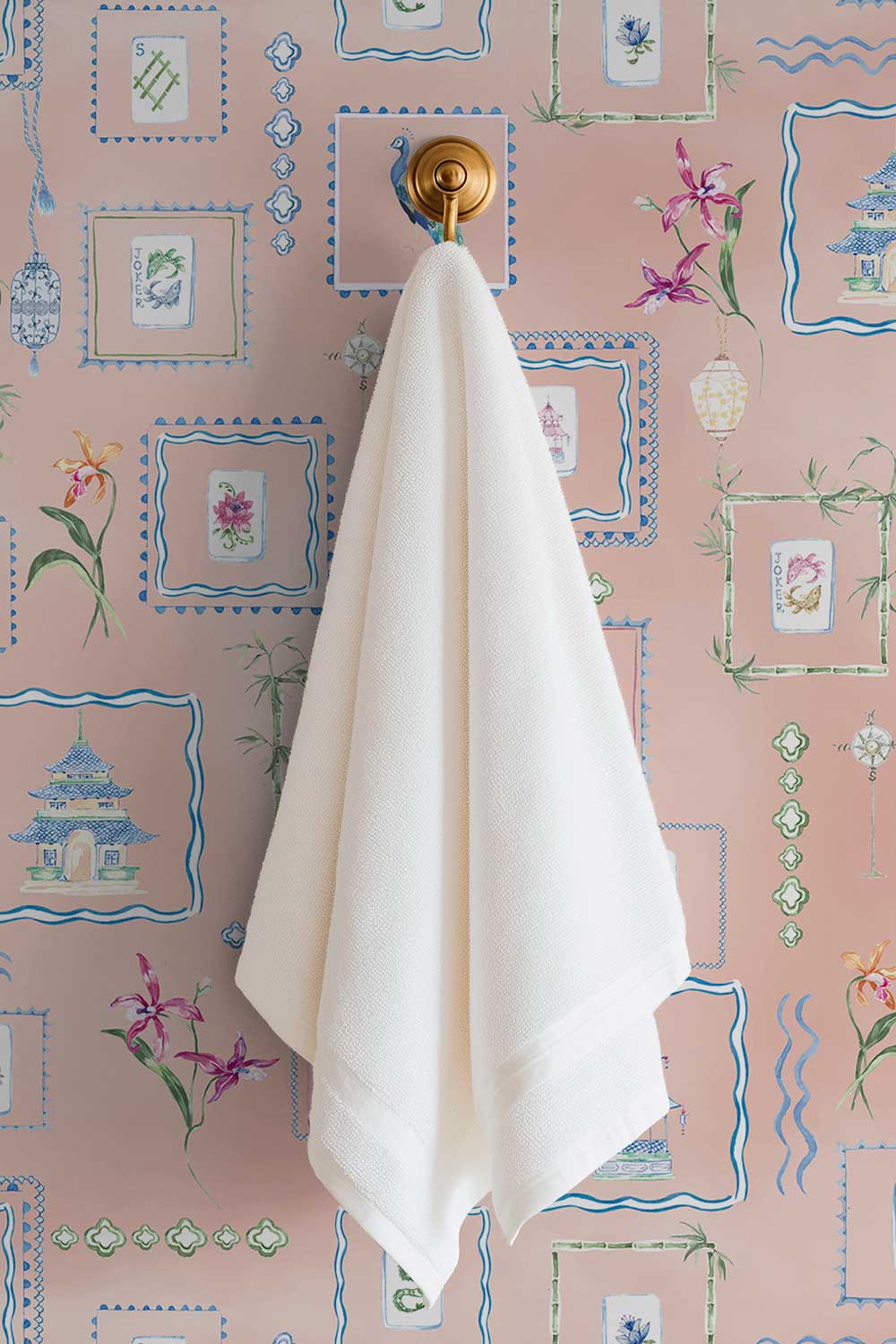A soft off-white cotton towel hangs from a round gold towel ring mounted on a pink wallpapered wall with blue and green botanical and frame motifs. Washable performance vinyl Mahjong Tiles wallpaper with hand-painted chinoiserie details, perfect for powder rooms, bathrooms, game rooms, and clubs.