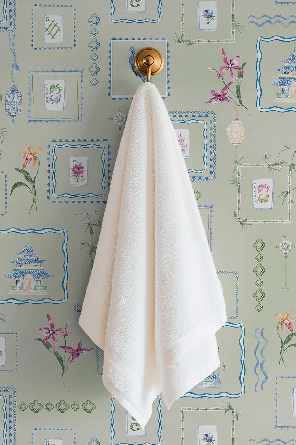 A white cotton hand towel hangs from a brass wall hook mounted on a sage green wallpaper with blue and pink botanical patterns. Washable performance vinyl Mahjong Tiles wallpaper with hand-painted chinoiserie details, perfect for powder rooms, bathrooms, game rooms, and clubs.