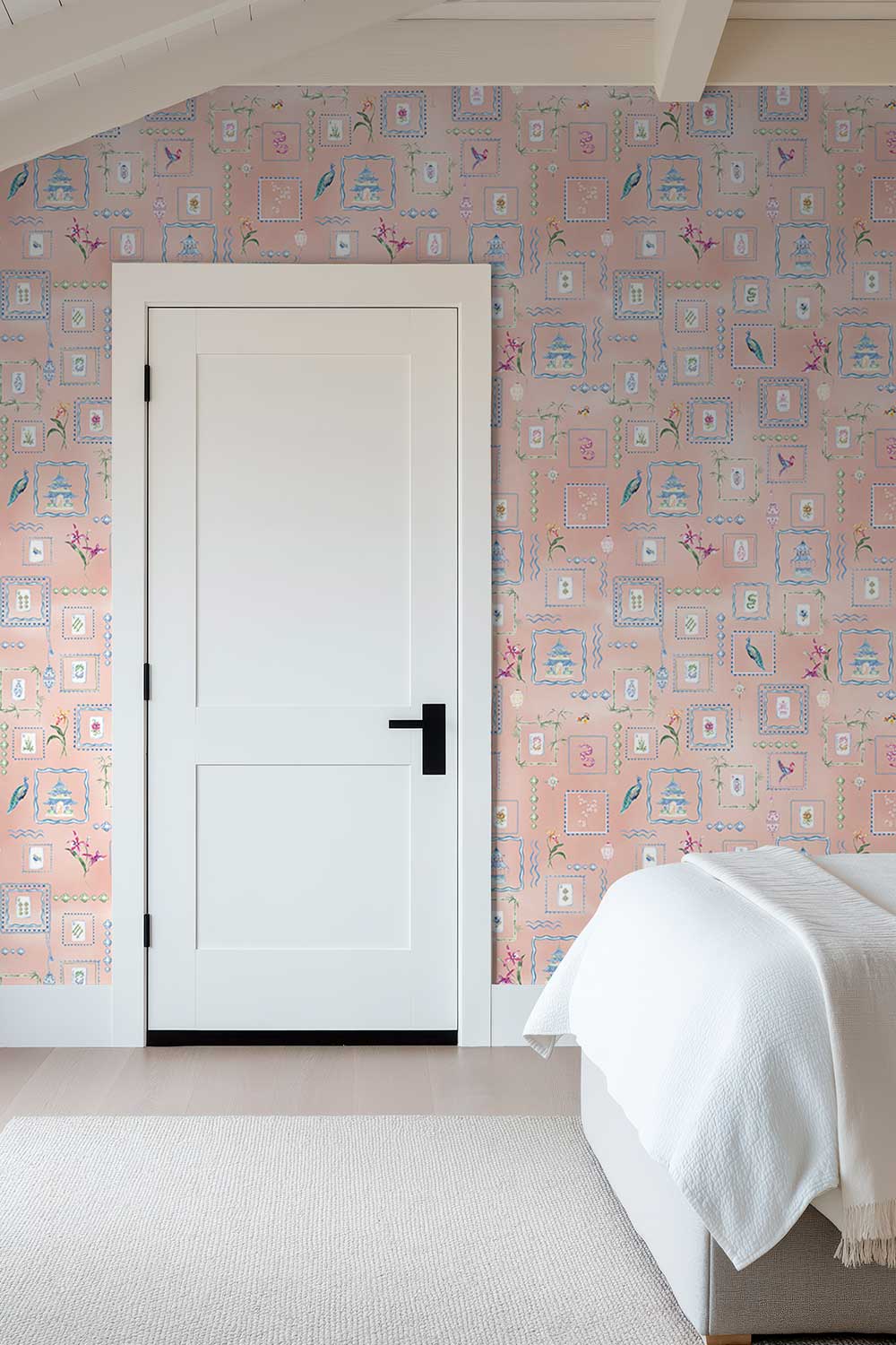 A white interior door with a black handle set in a bedroom with pink patterned wallpaper, a white bed with a white throw blanket, and a light rug. Soft pink coral Mahjong Tiles traditional matte wallpaper featuring hand-painted pastel watercolor mahjong tiles, pagodas, bamboo, ginger jars, birds, lanterns, and floral chinoiserie details.
