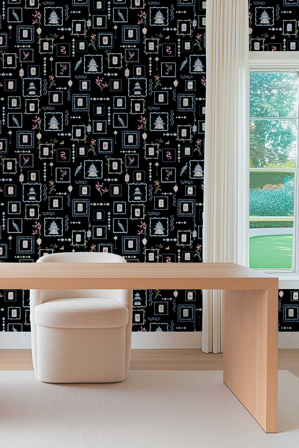 A styled interior room showing a black wallpaper with a repeating white, gray, and pink architectural pattern, paired with a light wood desk and a cream upholstered stool in front of a window. Black Mahjong Tiles traditional matte wallpaper featuring hand-painted pastel watercolor mahjong tiles, pagodas, bamboo, ginger jars, birds, lanterns, and floral chinoiserie details.