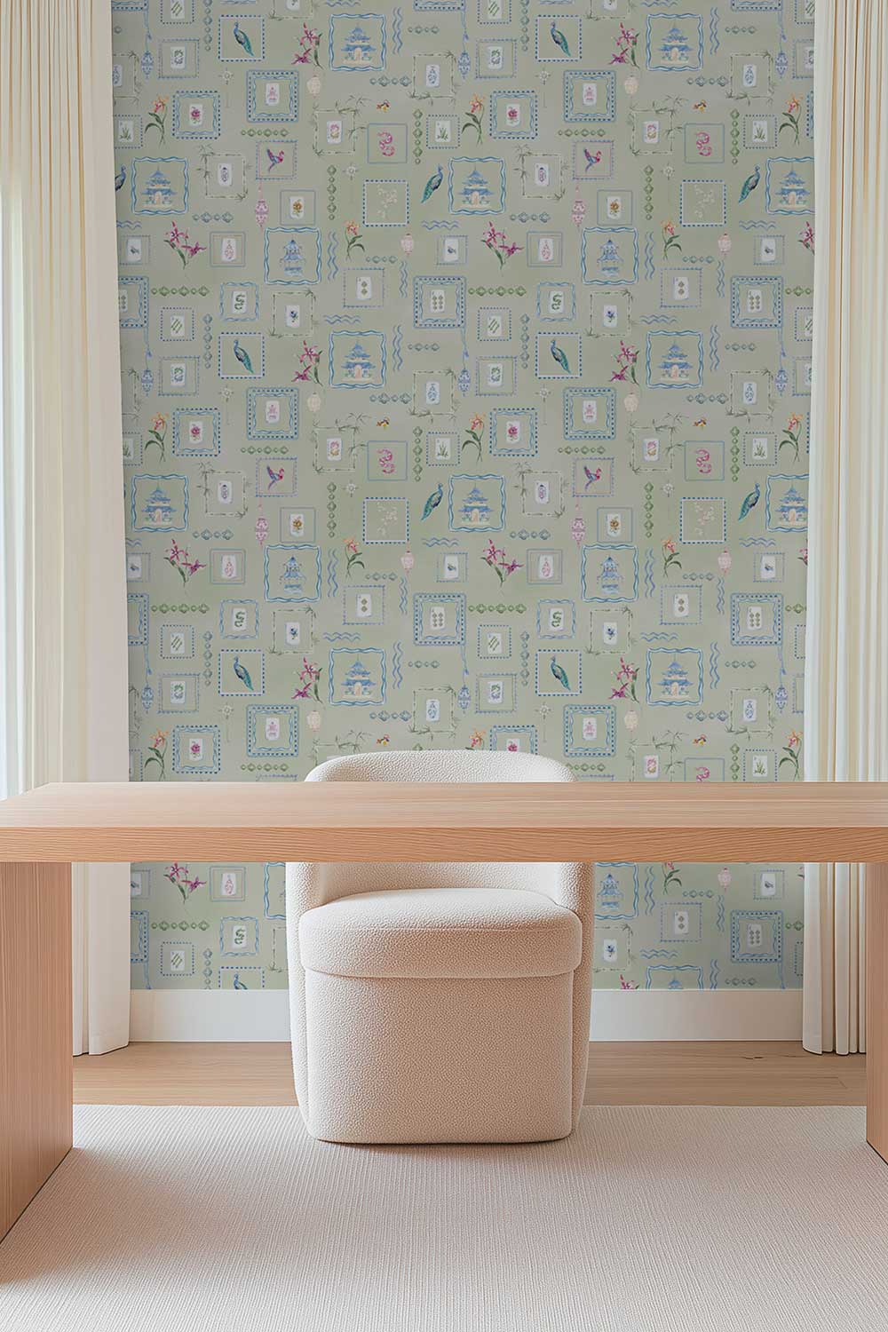 A styled interior scene showing a pale blue wallpaper with a repeating pattern of small blue, pink, and white decorative frames and motifs, behind a light wood desk and a cream upholstered stool. Soft green Mahjong Tiles traditional matte wallpaper featuring hand-painted pastel watercolor mahjong tiles, pagodas, bamboo, ginger jars, birds, lanterns, and floral chinoiserie details.