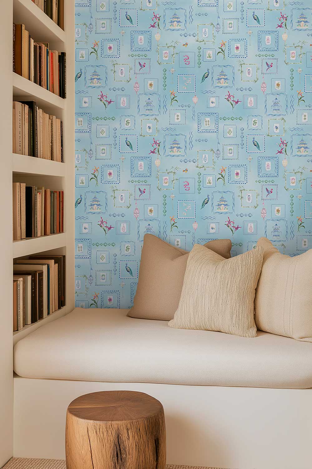 A cozy interior corner with a built-in bench seat, cream cushions, a small round wooden stool, and a light blue patterned wallpapered wall. Open bookshelves filled with books line the left side. Hand-painted watercolor Mahjong Tiles wallpaper featuring mahjong tiles, pagodas, bamboo, birds, ginger jars, lanterns, and chinoiserie artwork on a soft blue background printed on an all-natural and eco-friendly grasscloth wall-covering.