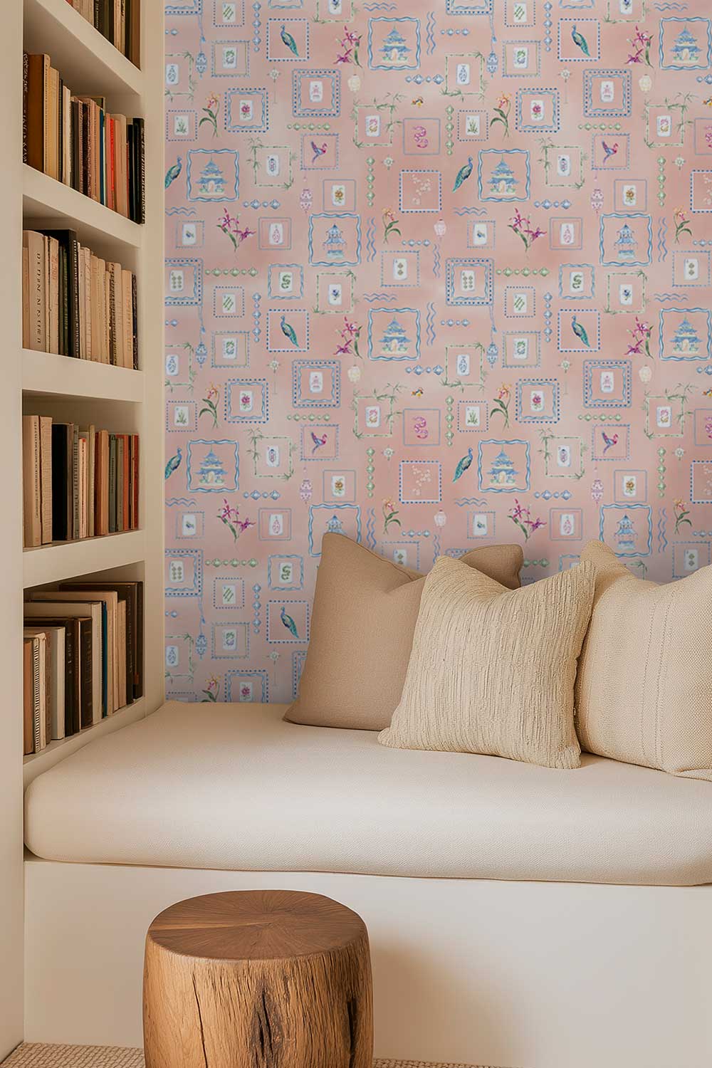A cozy interior corner with a built-in bench, cream cushions, a wooden stool, and a pink patterned wallpaper featuring small framed illustrations. A bookshelf with books is visible on the left. Soft pink coral Mahjong Tiles traditional matte wallpaper featuring hand-painted pastel watercolor mahjong tiles, pagodas, bamboo, ginger jars, birds, lanterns, and floral chinoiserie details.