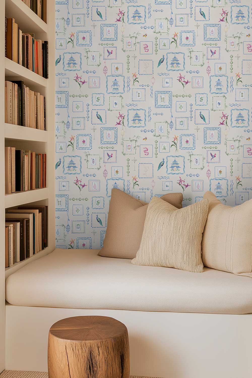 A cozy interior corner with a cream upholstered bench or daybed against a wall covered in a light blue and white patterned wallpaper featuring small framed illustrations and birds. Beige pillows rest on the bench, and a wooden stool sits in front. Off-white Mahjong Tiles traditional matte wallpaper featuring hand-painted pastel watercolor mahjong tiles, pagodas, bamboo, ginger jars, birds, lanterns, and floral chinoiserie details.