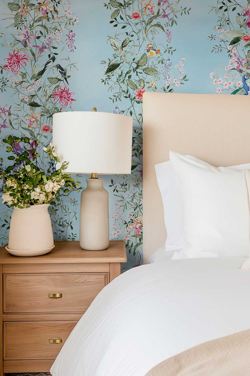 A styled bedroom corner featuring a light wood nightstand with a cream ceramic table lamp and a vase of flowers, next to a bed with white linens and a beige upholstered headboard, all set against a light blue wallpaper with a colorful floral and bird pattern. Natural grasscloth wallpaper featuring hand-painted Mahjong Garden Stripe with chinoiserie florals, peacocks, birds, bamboo, and botanical watercolor artwork.