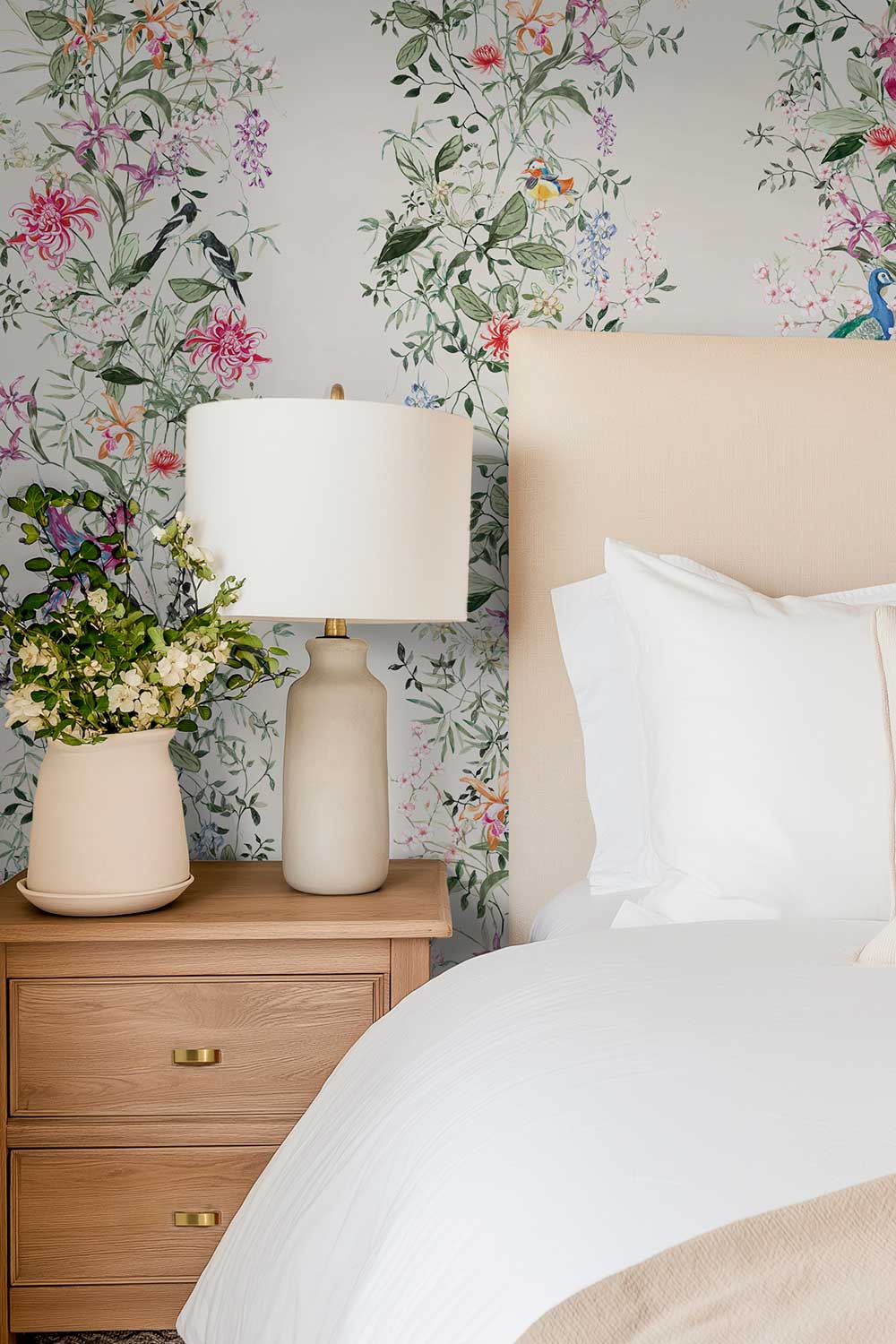 A styled bedroom corner featuring a floral and bird patterned wallpaper, a light wood nightstand with a cream ceramic table lamp and a potted plant, and a bed with white bedding and a beige upholstered headboard. Hand-painted Mahjong Garden Stripe wallpaper featuring watercolor chinoiserie florals, peacocks, birds, bamboo, and botanical stripes on an off-white background.
