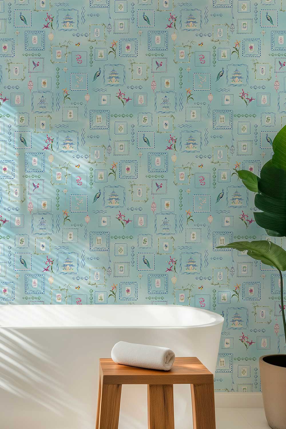 A bathroom scene featuring light blue wallpaper with a delicate vintage floral and architectural pattern, a white freestanding bathtub, a small wooden stool with a rolled white towel, and a large green potted plant. Washable performance vinyl Mahjong Tiles wallpaper with hand-painted chinoiserie details, perfect for powder rooms, bathrooms, game rooms, and clubs.
