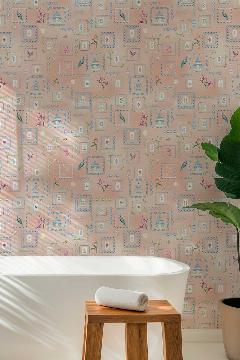 A bathroom scene featuring a light pink wallpaper with a repeating pattern of blue and green decorative frames, a white bathtub, a small wooden stool with a rolled white towel, and a large green plant on the right. Washable performance vinyl Mahjong Tiles wallpaper with hand-painted chinoiserie details, perfect for powder rooms, bathrooms, game rooms, and clubs.